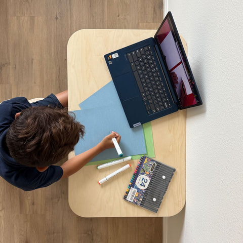 Child working creatively with markers and laptop on a Kids Table, surrounded by colorful paper.