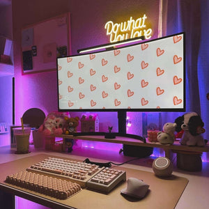 Decorated workspace featuring a monitor on a stand, illuminated keyboard, and motivational sign with pink lighting and heart-patterned screen.