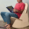 Child reading a book while seated on a Mini Rocker wooden kids rocking chair in a cozy, indoor setting.