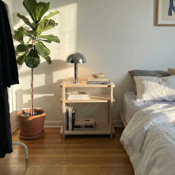 Modern Nightstand with books, lamp, and plant beside a cozy bed in a sunlit bedroom.