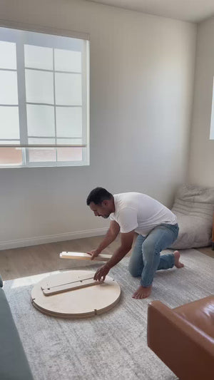 Man assembling a round coffee table on a rug in a bright living room, showcasing easy setup and stylish design.